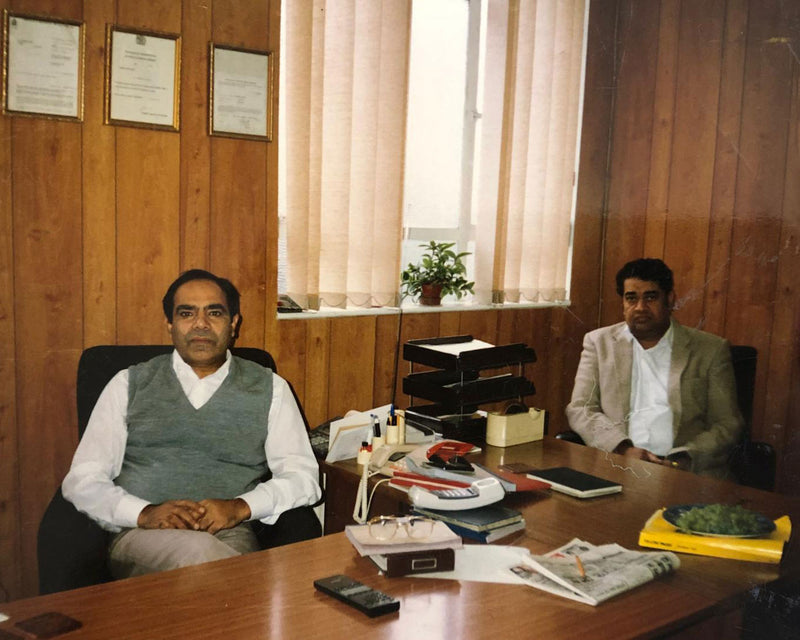 Two men seated at a wooden office desk with certificates, plants, and office supplies in a wood-paneled room.